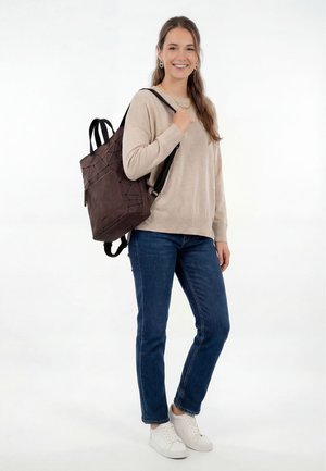 Lachende vrouw met een beige trui, blauwe jeans, witte sneakers, en een bruine rugzak met een geometrisch patroon over één schouder.