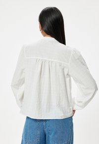 Camicia bianca a maniche lunghe con un motivo testurizzato sottile, schiena arricciata e colletto con bottoni, abbinata a jeans blu a gamba larga.
