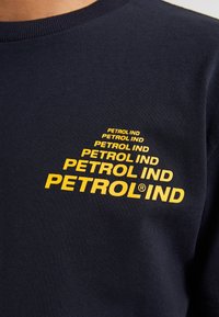 Námořnická modrá bavlněná tričko s žlutým nápisem "PETROL IND" v vrstveném, tučném písmu. Hladký kulatý výstřih a měkká textura.
