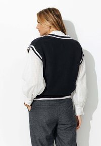 Gilet en maille marine à rayures blanches sur une blouse blanche texturée, associé à un pantalon à carreaux gris. Comprend un tissu superposé et une coupe décontractée.