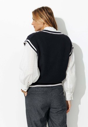 Gilet en maille marine à rayures blanches sur une blouse blanche texturée, associé à un pantalon à carreaux gris. Comprend un tissu superposé et une coupe décontractée.