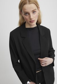 Zwarte blazer met een geknipte kraag, enkele knoopsluiting en voorkruiszakken. Draagt over een aansluitende zwarte top van gestructureerde stof.