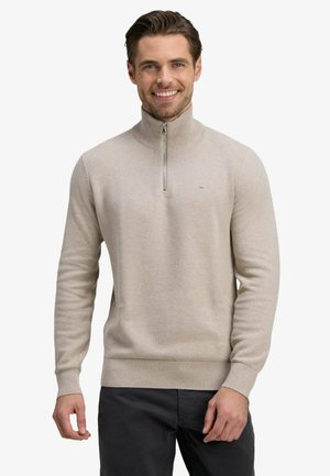 Homme souriant portant un pull beige à demi-fermeture éclair et un pantalon foncé, debout devant un fond blanc uni.