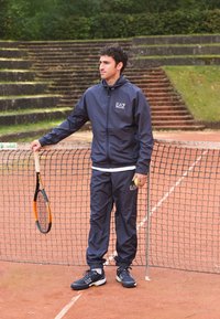 Navy athletic jacket and pants with EA7 logo, paired with black sneakers. The player holds a tennis racket beside a net on a clay court.