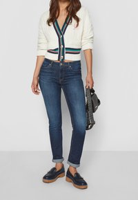 Cárdigan blanco de punto trenzado con cuello en V, ribete de rayas multicolores, combinado con jeans oscuros de corte ajustado y mocasines azul marino, sosteniendo un bolso con estampado.