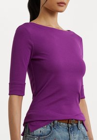 Purple prilegajoča majica s tremi četrtinami rokavov, izdelana iz gladkega blaga. Ima okroglo izrezovanje in stranske razpoke na robu.