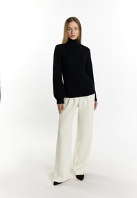 Maglione nero a coste con collo alto abbinato a pantaloni bianchi a gamba larga. La modella indossa décolleté nere a punta, mettendo in risalto le linee pulite dell'outfit.