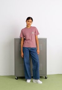 T-shirt di cotone rosa con logo sulla tasca, abbinata a jeans in denim blu larghi e sneakers bianche, su uno sfondo di armadietti grigi.