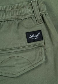 Pantalones de algodón verdes con un bolsillo trasero que tiene una etiqueta negra "Reell" y costuras en contraste a lo largo de las costuras.