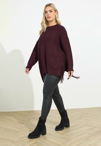 Burgundowa dziana bluza z bocznymi frędzlami, zestawiona z czarnymi skórzanymi legginsami i czarnymi botkami do kostek, na jasnym tle.