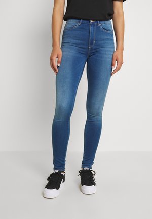 Personne portant un jean skinny bleu, un haut noir et des baskets noires et blanches, debout devant un fond clair uni.