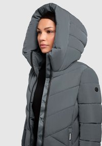Kvinde iført en fyldig grå hættejakke med diagonal quiltning og sort lynlås over en sort top, der står til venstre på en ensfarvet baggrund.