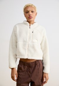 Kurze, cremefarbene Fleecejacke mit Reißverschluss, hohem Kragen und einer Fronttasche. Weiche Textur, verkürzter Schnitt und lässige Passform.