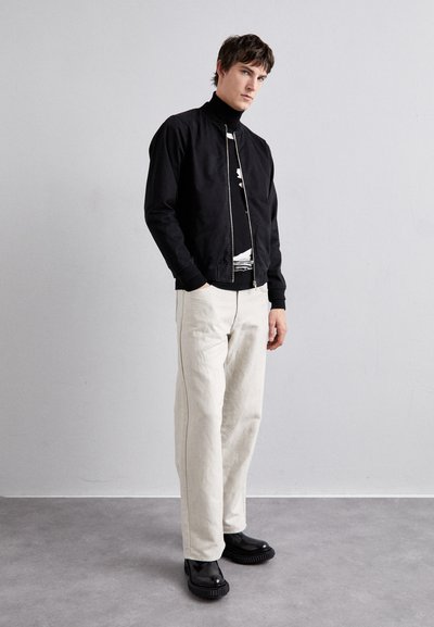 Chaqueta bomber negra con cierre de cremallera, llevada sobre una camiseta gráfica en blanco y negro, combinada con pantalones de color claro y zapatos negros gruesos.