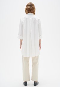 Balta krekli ar garām piedurknēm un krokotu aizmuguri, apvienots ar gaiši bežām plašām biksēm un melniem slīdošajiem apavos. Vienkāršs un tīrs dizains.