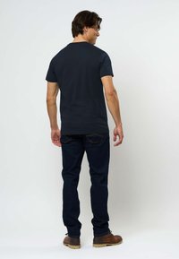 Muškarac stoji okrenut leđima, obučen u tamnoplavu majicu, tamnoplave traperice i smeđe kožne cipele, na jednobojnoj svijetloj pozadini.