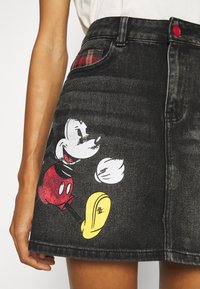 Black denim skirt featuring a Mickey Mouse graphic in red and yellow. It has a plaid accent in the back pocket and a pink button.