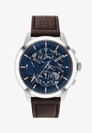 Navy blue watch with a skeleton dial, silver stainless steel case, and brown leather strap. Features date and day indicators, chronograph sub-dials.