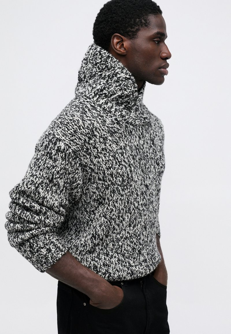 Maglione lavorato a maglia in bianco e nero con colletto alto, caratterizzato da un motivo testurizzato e una vestibilità comoda, abbinato a pantaloni scuri.