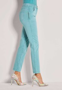 Frau, die schmal geschnittene türkisfarbene Hose mit seitlichem Reißverschlusstasche und mehrfarbig gemusterten High Heels trägt, steht seitlich.