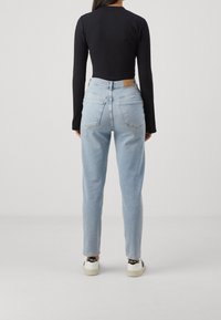 Kvinna i ljusblå högmidjade jeans, svart ribbad långärmad topp och vita sneakers med svarta detaljer, stående med ryggen vänd.