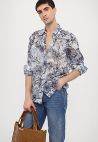 Camicia a maniche corte, con bottoni, dal motivo floreale blu chiaro e blu scuro; indossata con jeans blu e tiene una borsa a tracolla marrone.