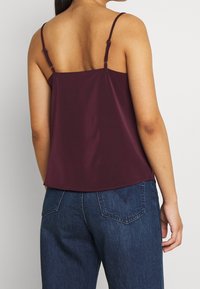 Camisola in satin color bordeaux con spalline sottili regolabili, caratterizzata da una vestibilità comoda e una trama liscia, abbinata a jeans in denim blu scuro.