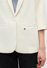 Μπλέιζερ σε κρεμ απόχρωση με γιακά notch, κλείσιμο με ένα κουμπί, δύο μπροστινές τσέπες, φορεμένο πάνω από γκρι τοπ και σκούρο παντελόνι.