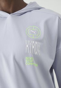 Világos lila kapucnis póló, kapucnival, fehér és neon zöld grafikával, beleértve egy logót és a "NEVER STOP COMPETING" feliratot.