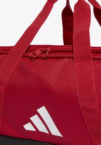 Bolsa de lona roja con un cierre resistente, tejido texturizado, correas anchas y un gran logo blanco en el lateral. Presenta un acabado suave.