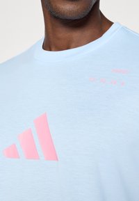Jasnoniebieska, krótko rękawowa koszulka z miękkiego materiału, z dużym różowym logo Adidas oraz tekstem na lewym górnym boku.