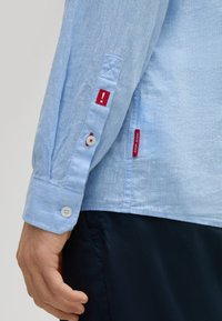 Camicia azzurra chiara a maniche lunghe, con dettagli rossi e bottoni bianchi. Il tessuto sembra leggero con una finitura texturizzata.