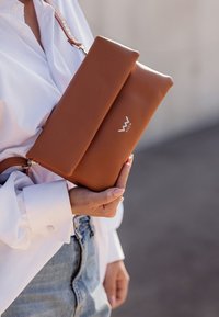 Kožená taška v barvě tan s hladkým povrchem, skládacím designem a stříbrným logem. Obsahuje popruh na přes rameno a zipové uzavření.