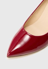 Escarpin rouge en cuir verni à bout pointu avec une finition lisse et brillante et une doublure intérieure beige. Il présente un talon fin et un design minimaliste.