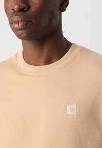 Sudadera beige de cuello redondo hecha de una tela suave, con una pequeña etiqueta de logo en relieve en la esquina inferior izquierda. Textura lisa, puños acanalados.