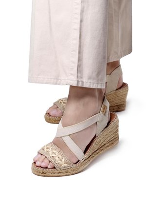 Beige espadrille wedge sandals with woven fabric upper, elastic cross straps, and jute-wrapped sole; foot partially visible.