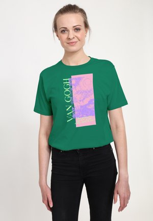 Kvinde med lyst hår bundet tilbage iført en grøn Van Gogh grafik t-shirt og sorte bukser, stående mod en ensfarvet hvid baggrund.