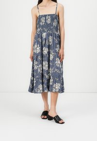 Blaues, floral bedrucktes Kleid mit Spaghetti-Trägern, gerafftem Oberteil und gestuftem Rock. Getragen mit schwarzen Blockabsatz-Mules.