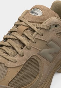 Κοντινό πλάνο μπεζ αθλητικού παπουτσιού New Balance με υφή από δίχτυ, επενδύσεις από σουέτ, κορδόνια που διασταυρώνονται και λογότυπο της μάρκας στην πλευρά και στη γλώσσα.