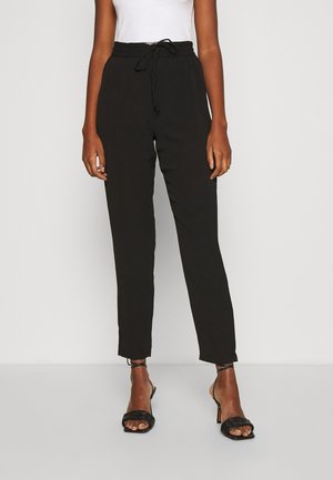 Pantalons noirs légers et fuselés avec une taille à cordon de serrage, présentant une texture lisse et un drapé subtil, associés à des escarpins noirs à lanières.