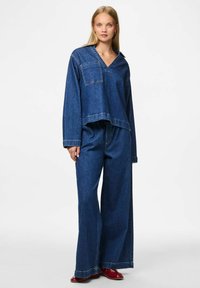 Denim apģērbs, kas ietver brīva piegriezuma zilu topu ar kabatu un platām zilu džinsu biksēm. Materiāls ir ar vidēju tekstūru un vieglu nodilumu.