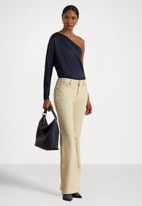 Σκούρο μπλε τοπ με ένα μανίκι σε συνδυασμό με κρεμ καμπάνες παντελόνι. Το μοντέλο κρατάει μία τσάντα σε σκούρο μπλε χρώμα. Το ντύσιμο διαθέτει απαλές υφές και διακριτικές μεταλλικές λεπτομέρειες.