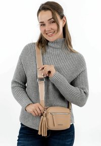 Borsa a tracolla beige in pelle morbida, con tasca con zip e dettaglio con pon-pon. Tracolla regolabile e forma arrotondata. Maglione in tessuto texture visibile.