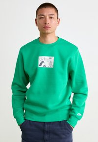 Sweatshirt in pile verde con scollo rotondo e polsini a coste. La grafica centrale presenta illustrazioni e testo, logo Nike a marchio sulla manica.