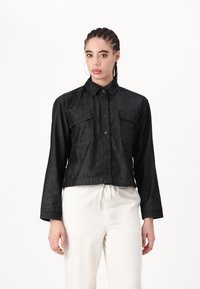 Chaqueta recortada de mezclilla negra con frente de botones, dos bolsillos en el pecho y mangas largas. Textura suave con detalles de costura mínimos.