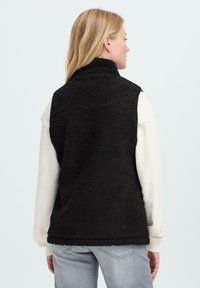 Vrouw met blond haar die een zwarte fleece vest draagt over een witte longsleeve shirt en lichtblauwe jeans, staand met haar rug naar de camera tegen een effen achtergrond.