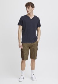 Marineblå v-hals t-shirt, olivengrøn cargo shorts med sidelommer, hvide sportsstrømper og sort-hvide sneakers. Ensfarvet baggrund.