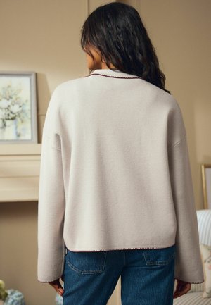Beiger Pullover mit langen Ärmeln, einem runden Ausschnitt und kontrastierendem roten Nähten am Saum. Getragen mit blauen Jeans.
