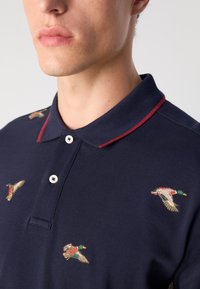 Marineblå polo-skjorte med rødstripet krage; har broderte andedesign i forskjellige farger; tre-knapp-stolpe; teksturert stoff.