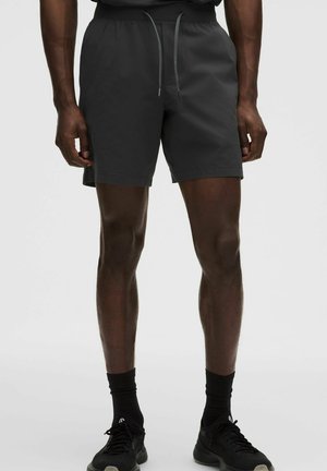 Mann trägt schwarze Kordelzug-Shorts, schwarze Socken und schwarze Sportschuhe, steht vor einfachem Hintergrund.